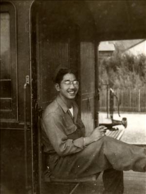 A young Asian man in army fatigues sits on a wooden bench holding a cigarette and smiling. 